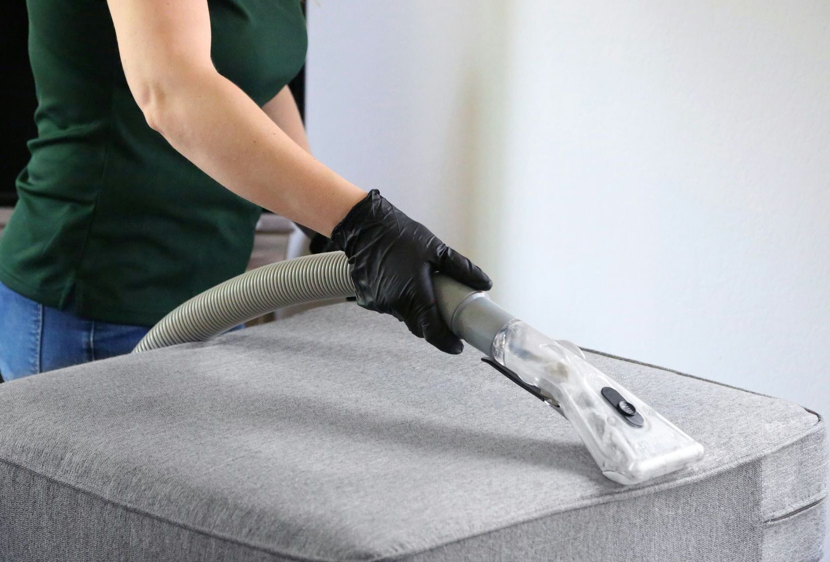 a woman in a green shirt and black gloves vacuuming a gray ottoman